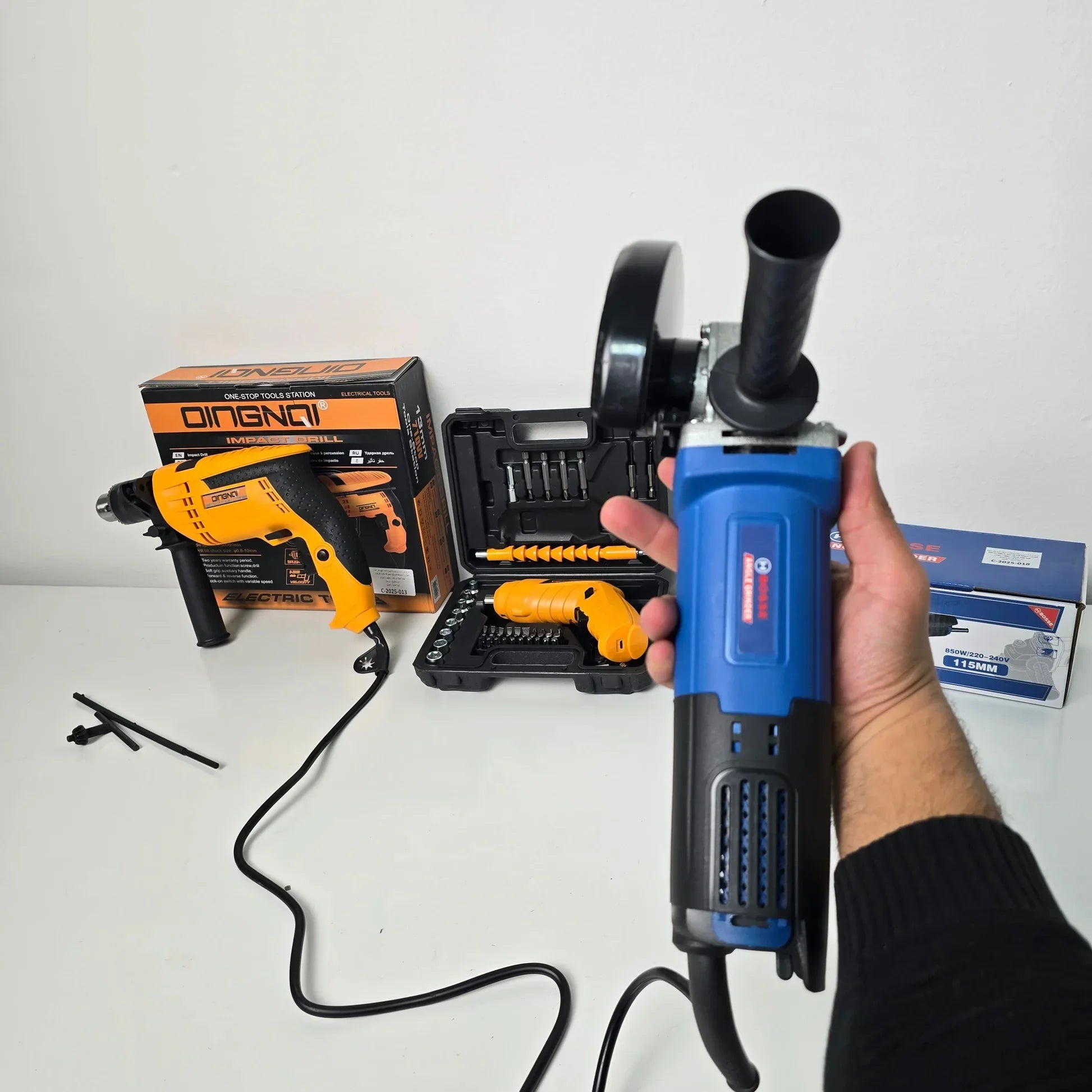 Hand holding a blue Bosch electric angle grinder with black handle, with an orange QINGQI impact drill and tool kit on white surface