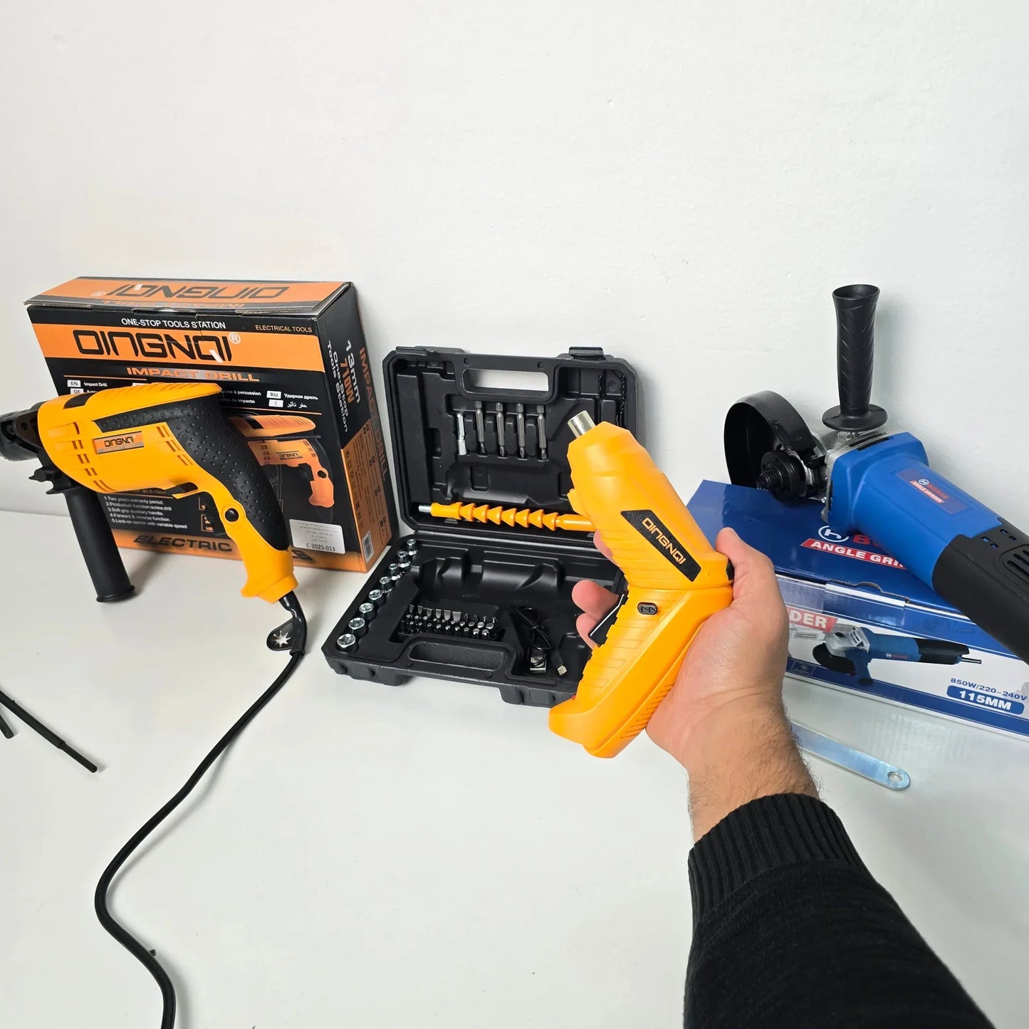 Hand holding yellow QINGNQ heat gun with impact drill and angle grinder tools on white surface